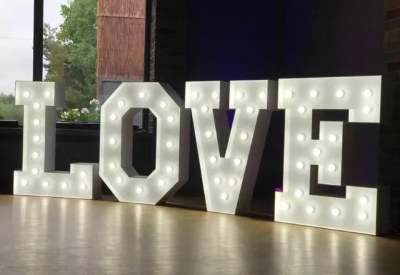 Podświetlany napis LOVE z białymi żarówkami stoi na drewnianej podłodze sali