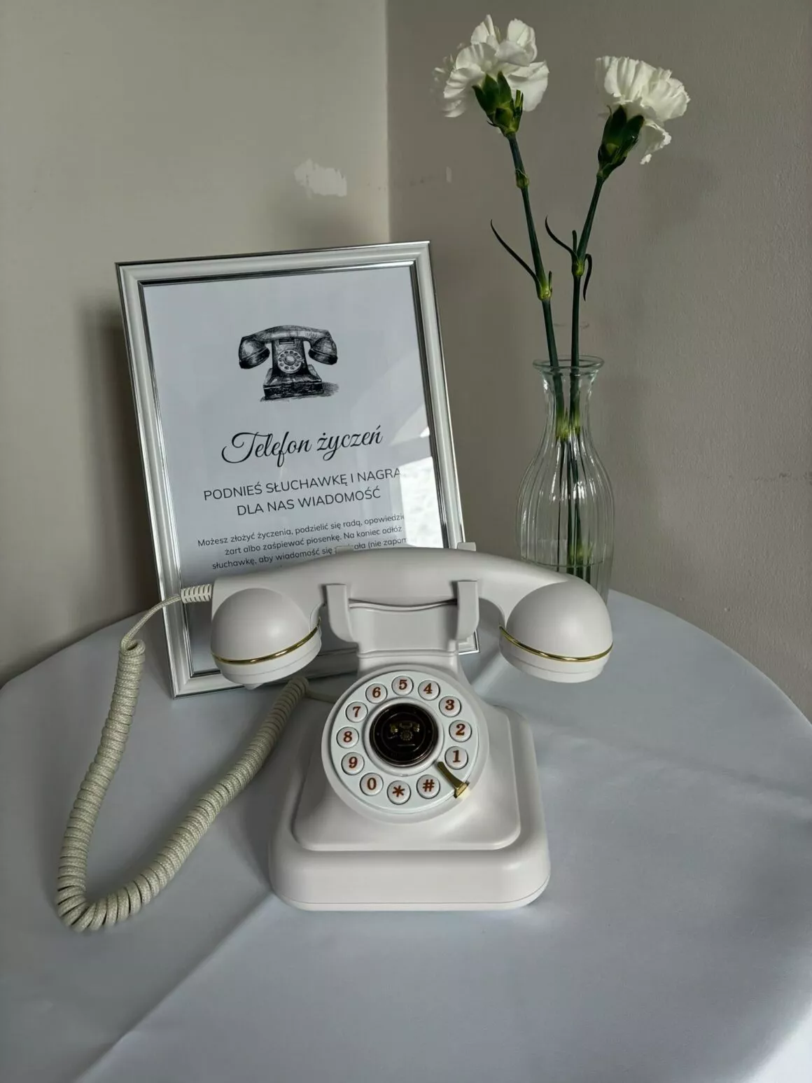 Biały telefon życzeń retro z tarczą na stoliku, obok ramka z instrukcją dla gości i białe goździki