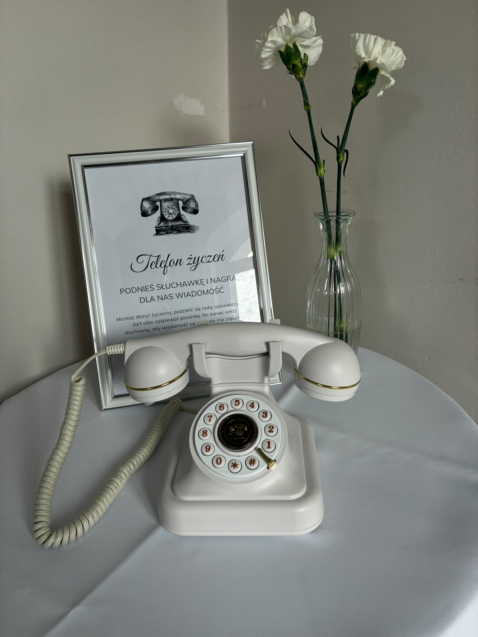 Biały telefon życzeń retro z tarczą na stoliku, obok ramka z instrukcją dla gości i białe goździki
